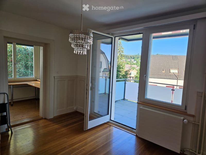 Bright room with wooden flooring, large windows, and a balcony access. Features a chandelier and a small table near the window.