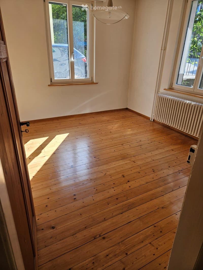 Empty room with wooden flooring, large windows, radiator, and a hanging light fixture.
