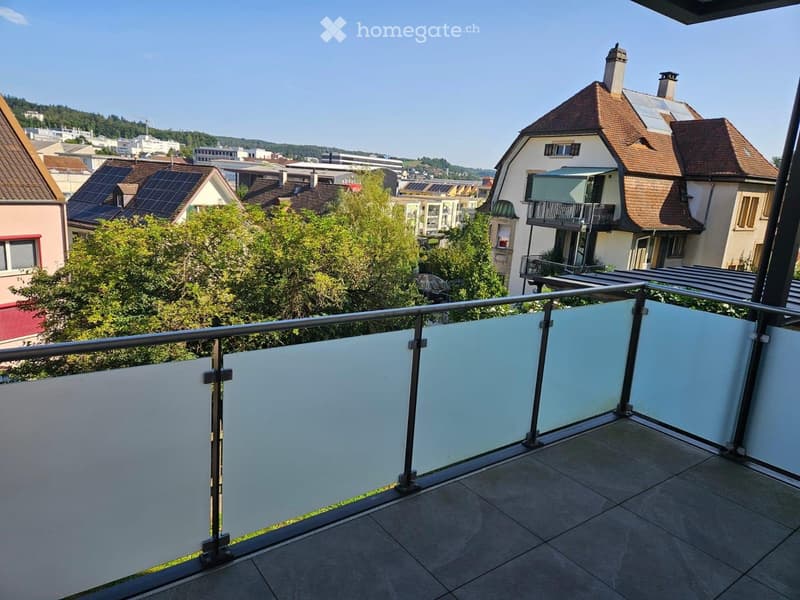 Balcony with modern glass railing, tiled floor, and scenic view of a residential neighborhood with houses, trees, and distant hills.