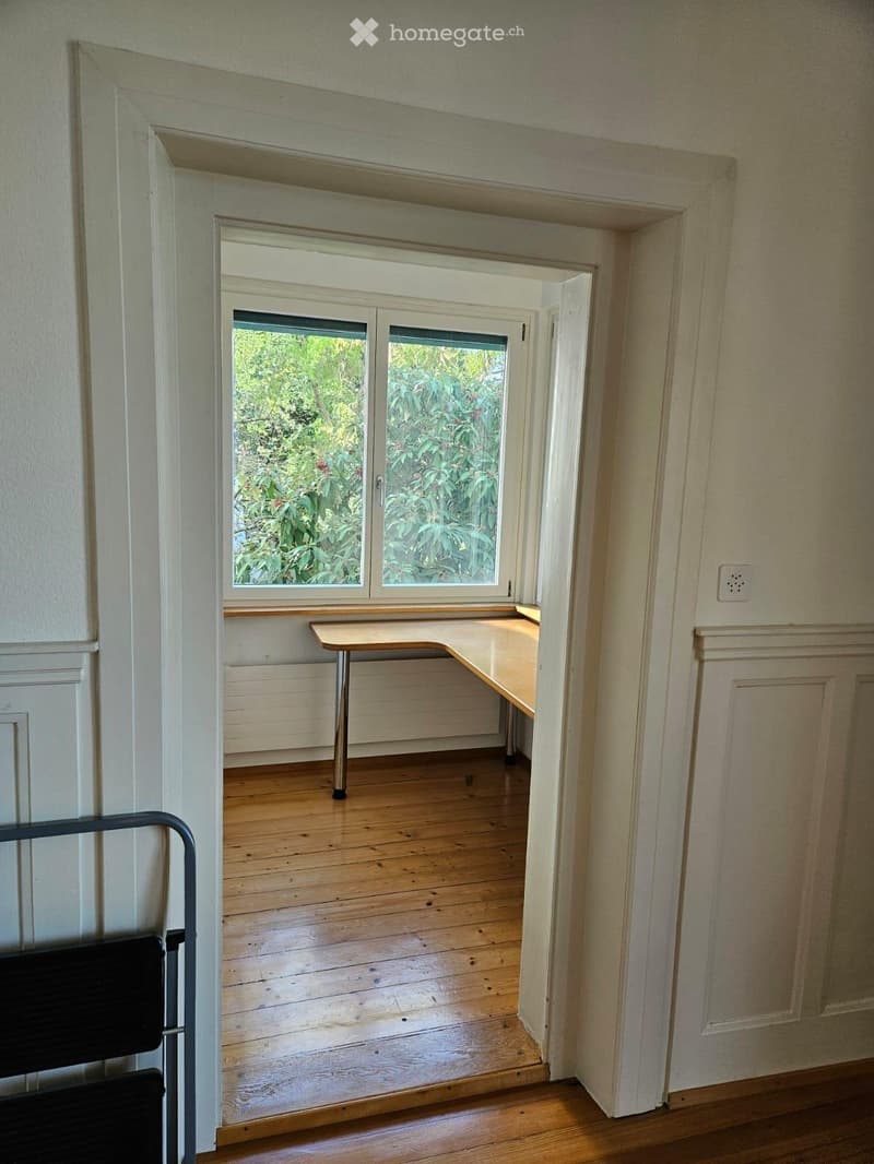 Empty room with wooden flooring, white walls, and a small table near a window with a view of greenery outside.