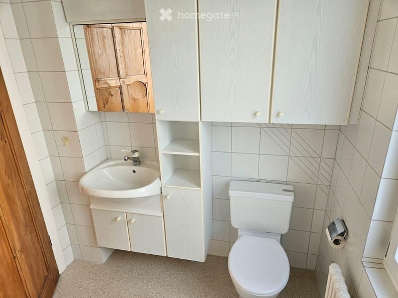 A small bathroom with a white sink, toilet, wooden cabinet, and mirror. The walls are tiled, and the floor is carpeted.