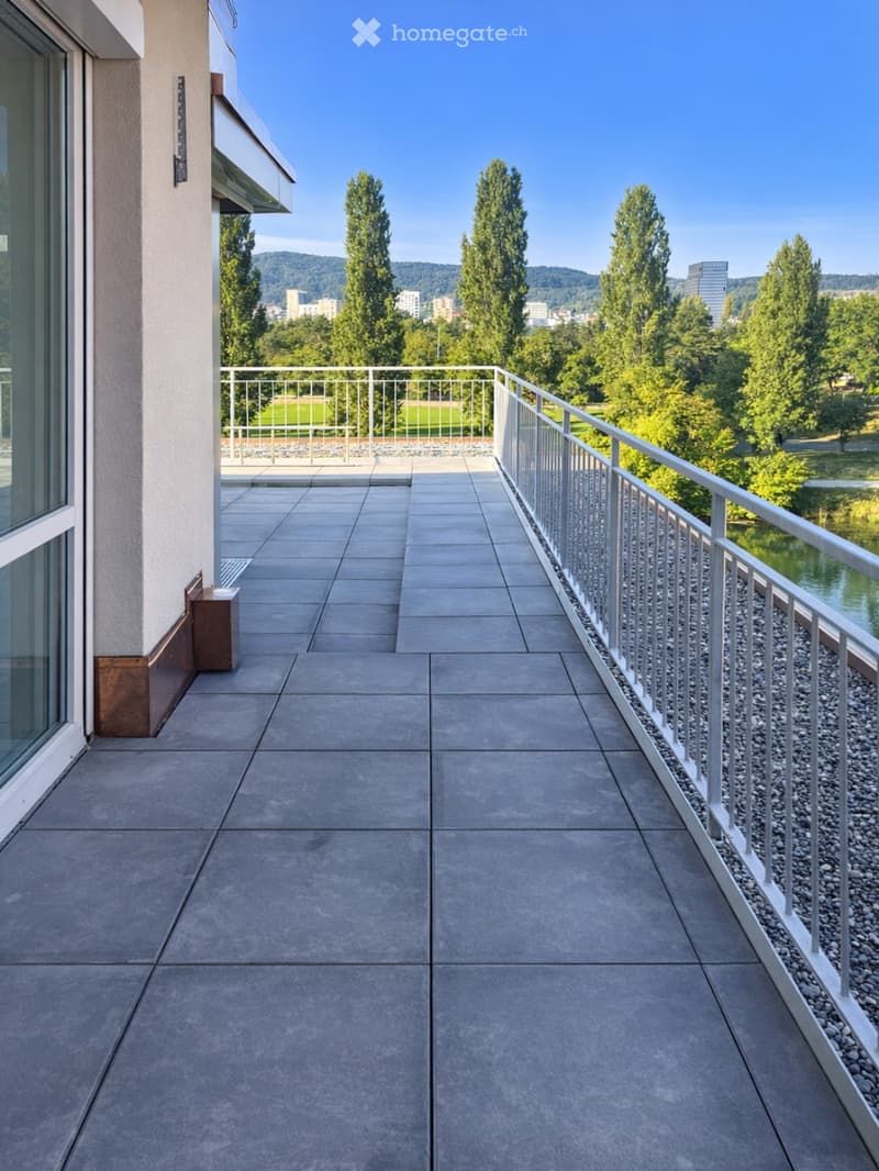 Modern balcony with grey tiled floor, white metal railing, and scenic city view with trees and water.
