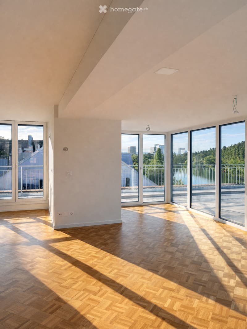 Bright, empty room with wooden herringbone flooring and large windows offering a scenic view of a lake and surrounding greenery.