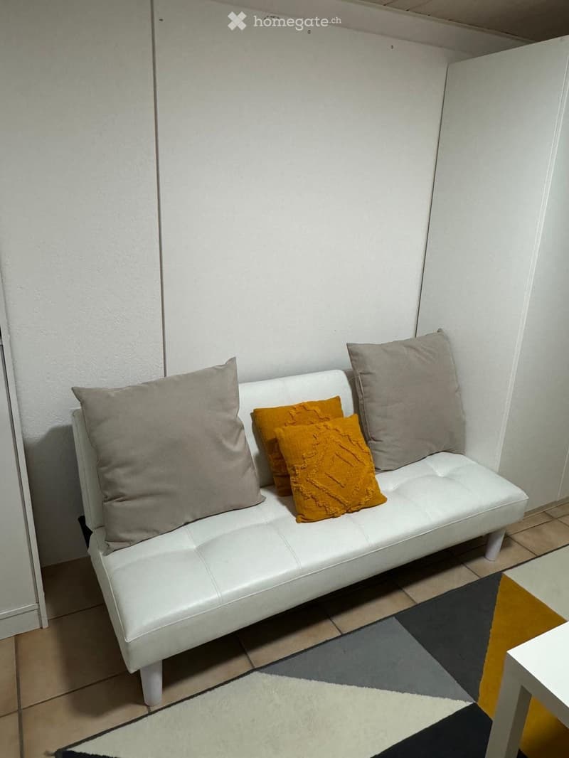 A white leather sofa with grey and orange pillows, placed in a corner of a room with tiled flooring and a small rug.