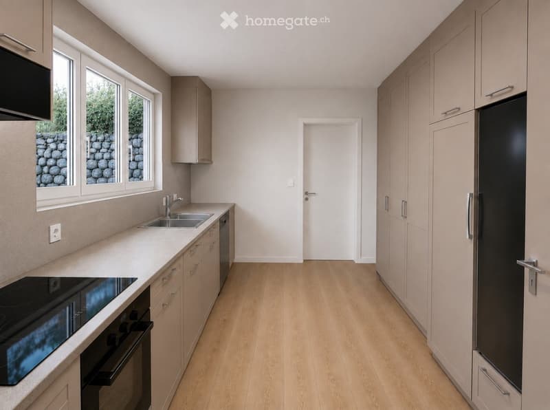 Modern kitchen with light wooden flooring, light-colored cabinets, and stainless steel appliances including a sink, oven, and refrigerator. The kitchen features a window with a view of a stone wall and greenery outside.