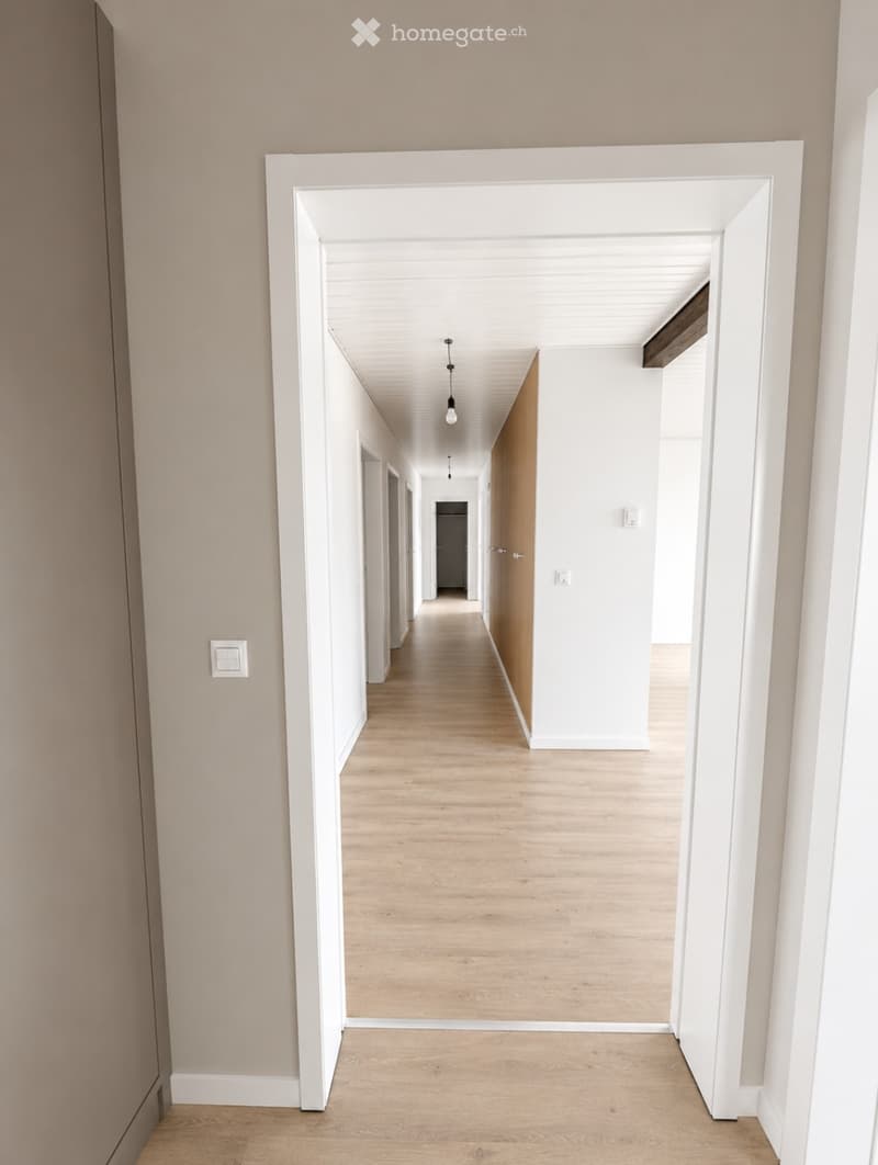 Modern hallway with light wooden flooring, white walls, and recessed lighting. Features white baseboards and a wooden door frame on one side.