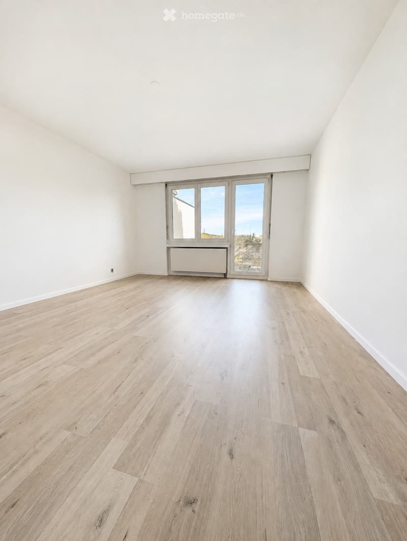Empty room with light wooden flooring, white walls, and a large window with a view of the outside.