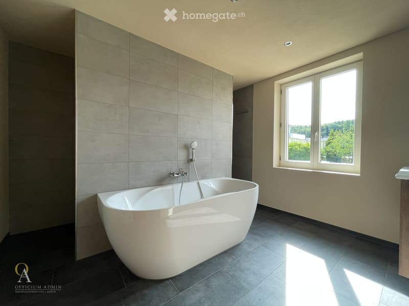 Modern bathroom with a large, freestanding bathtub, sleek grey tiles, and a large window offering a view of greenery outside.