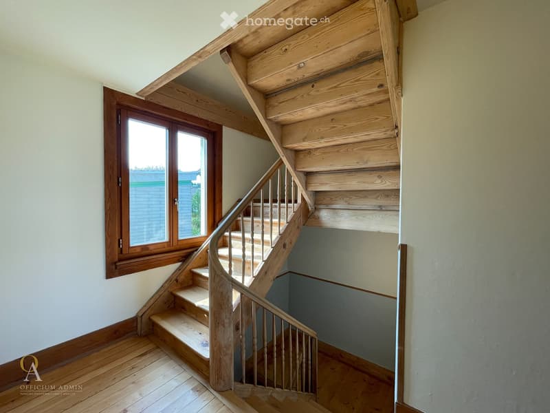 Wooden staircase with a curved handrail and wooden balusters, leading to an upper level. The staircase is flanked by light-colored walls and a window on one side, allowing natural light to enter.