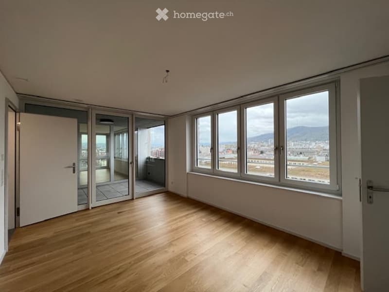 Empty room with wooden flooring, large windows offering a city view, and a sliding glass door leading to a balcony.