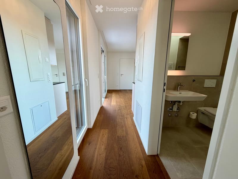 A narrow hallway with wooden flooring, white walls, and doors leading to various rooms, including a bathroom with a sink and toilet.