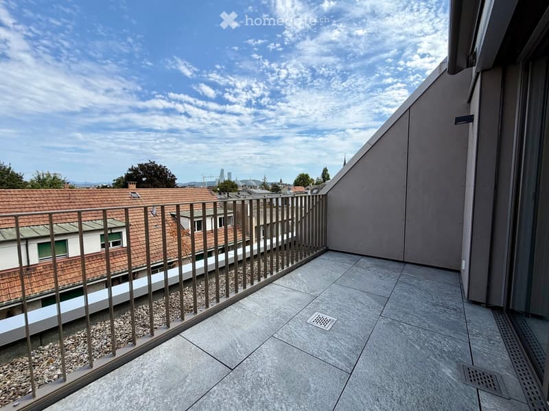 Balcony with grey tiled floor, metal railing, and a view of rooftops and cityscape under a partly cloudy sky.