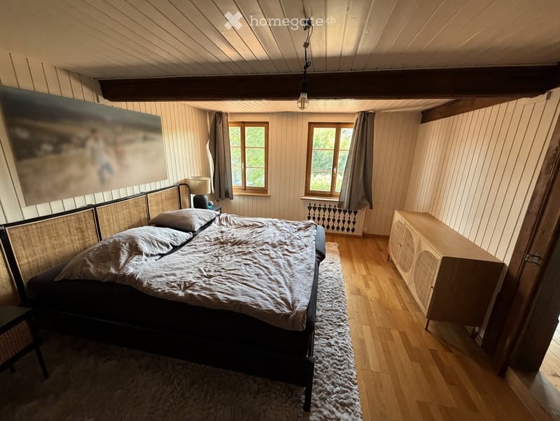 Cozy bedroom with wooden floors, a large bed with a woven headboard, nightstand, and a wooden cabinet. Natural light from two windows with curtains, and a painting on the wall.