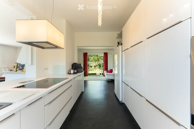 Modern kitchen with white cabinets, a central island, and an induction cooktop. A range hood is mounted above the island. The kitchen opens to a living area with a white sofa, ottoman, and a view of a garden through large windows with red curtains.