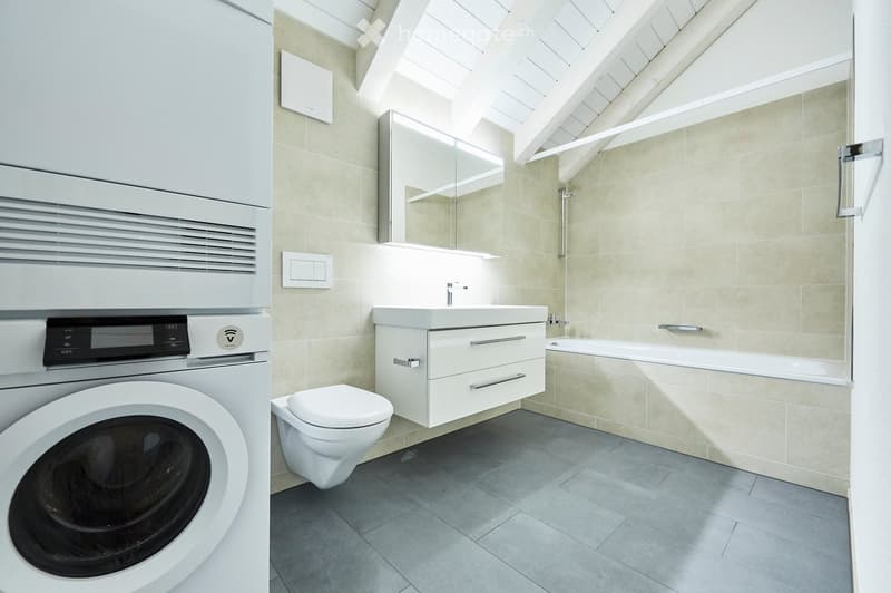 Modern bathroom with a toilet, sink, bathtub, mirror, and washing machine. The space features light-colored tiles and a sleek, contemporary design.
