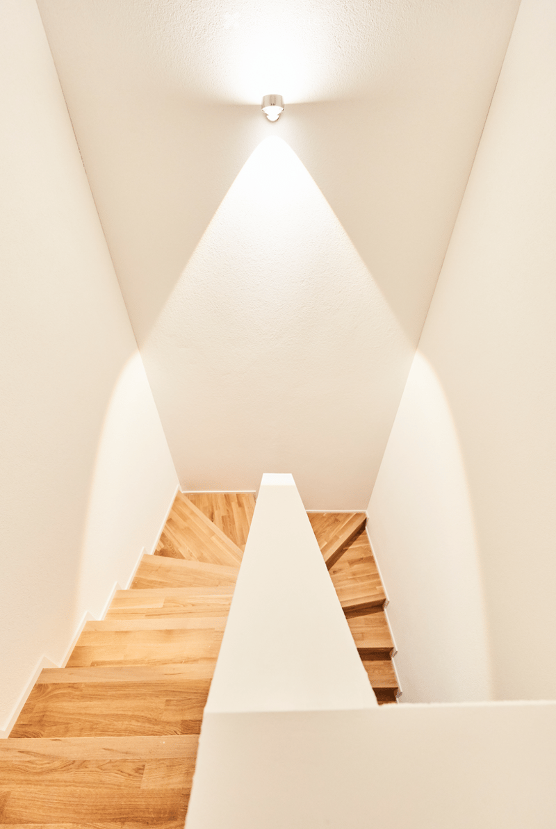 Modern, minimalist staircase with wooden steps and white walls, illuminated by a single ceiling light casting a triangular pattern of light on the wall.