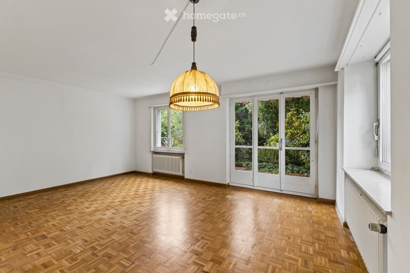 Empty room with wooden herringbone flooring, white walls, large windows, and a hanging pendant light fixture.