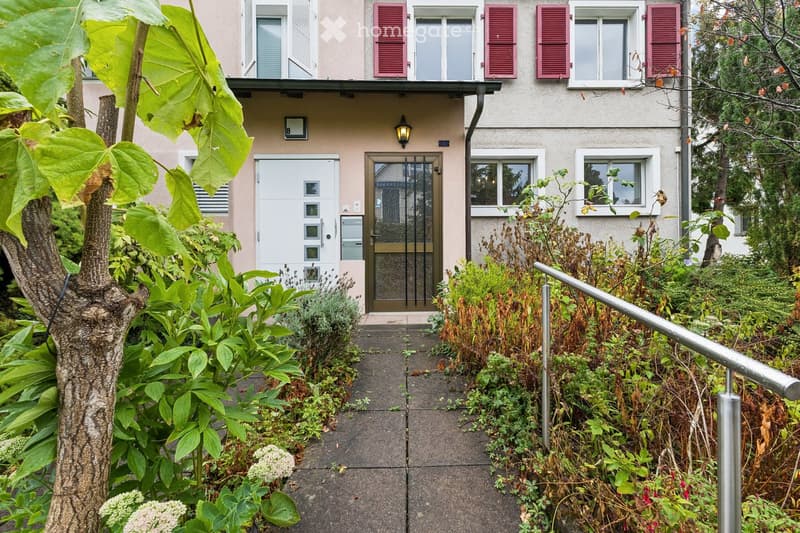 The image shows a garden area leading to the entrance of a two-story building. The pathway is bordered by lush greenery, including shrubs and a tree with large leaves. The building features a white door with a glass panel, a small porch light, and multipl