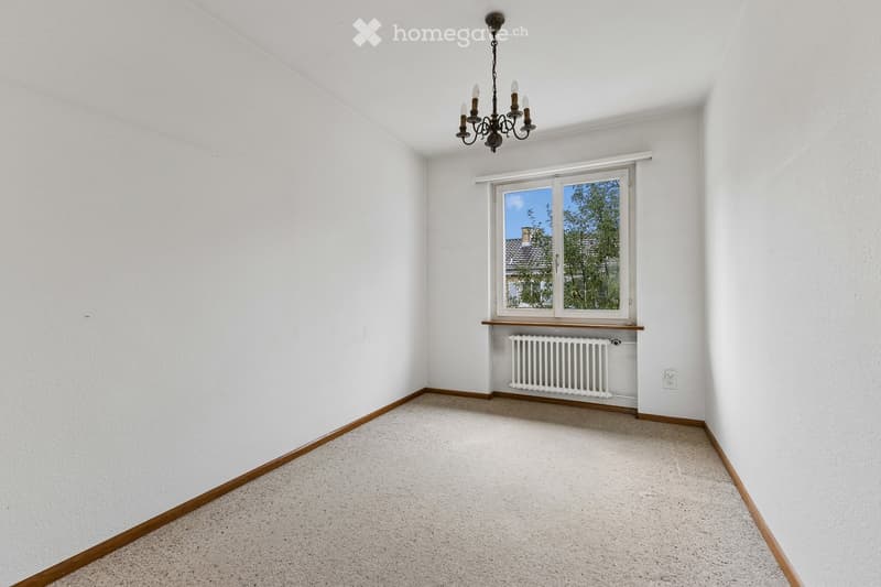 Empty room with white walls, carpeted floor, a window with a view of trees, a radiator, and a chandelier.