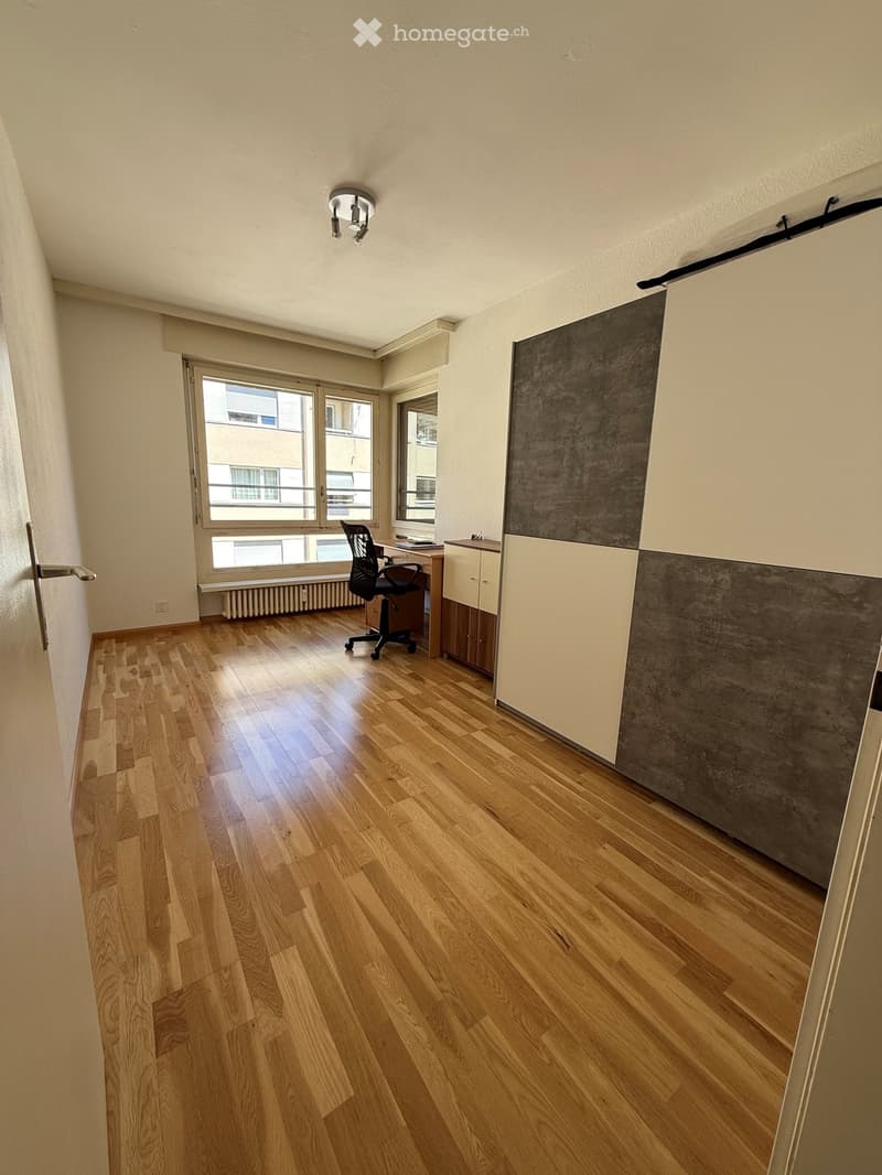 A modern, well-lit room with wooden flooring, a large window, a black office chair, a wooden desk, and a cabinet with a grey and white design.