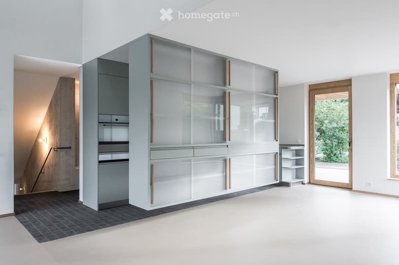Modern, minimalist kitchen with sleek, grey cabinetry and glass panels, featuring a built-in oven and microwave, and a small open shelving unit near the window.