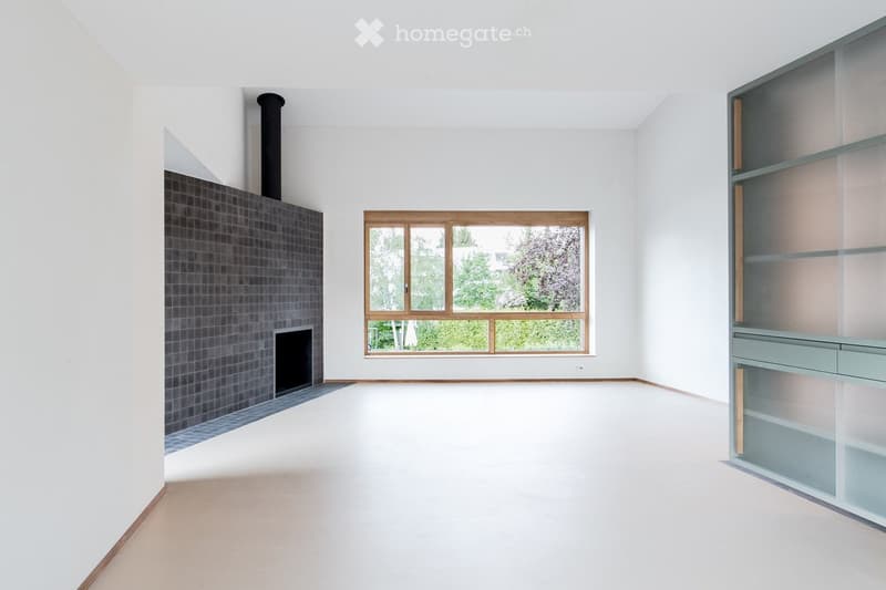 Completely empty room with white walls, wooden floor, fireplace, and large window with wooden frame.