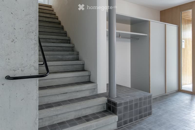 Modern concrete staircase with metal railing leading to an upper level, featuring a built-in shelf unit and a wooden door.