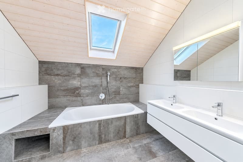 Modern bathroom with a large skylight, white cabinets, grey tiled walls, a bathtub, and a double sink with modern faucets.