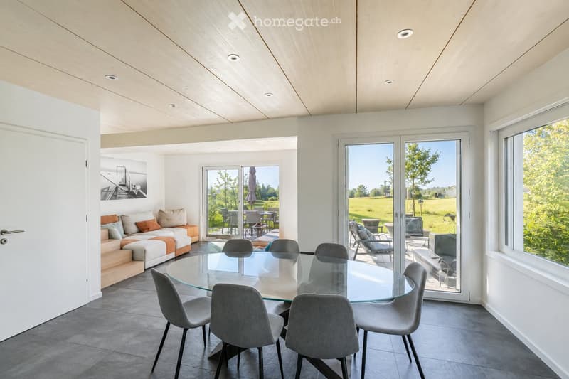 Modern living and dining area with a white sofa, glass dining table, and grey chairs. Large windows and sliding glass doors open to an outdoor patio with additional seating.