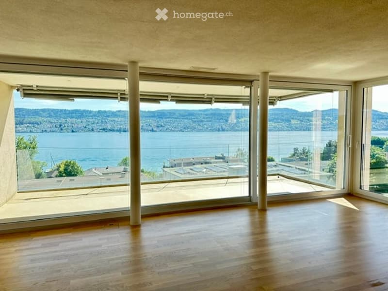 Bright, empty room with large glass walls offering a scenic view of a body of water and distant hills. The floor is made of polished wood, and the space is well-lit with natural light.