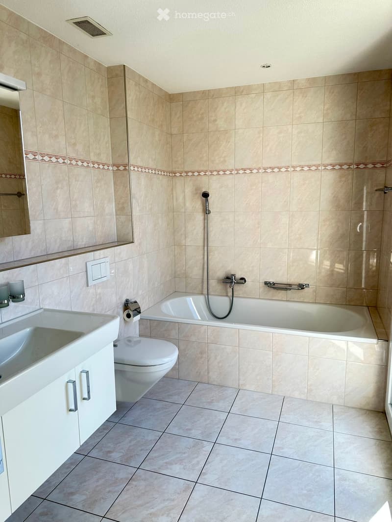 Modern bathroom with beige tiles, a white sink, toilet, bathtub, and a mirror above the sink. Features include a handheld showerhead, a towel rack, and a light switch.
