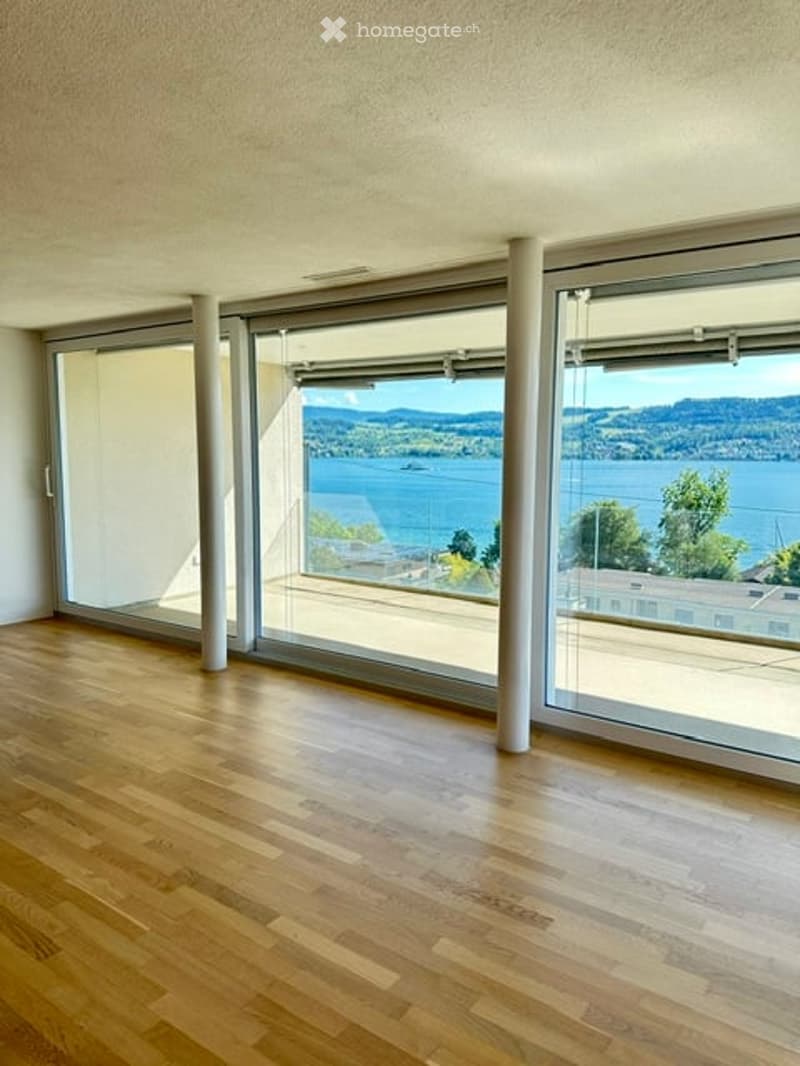 Bright, empty room with large floor-to-ceiling windows offering a scenic view of a lake and mountains. The room features a polished wooden floor and white walls.