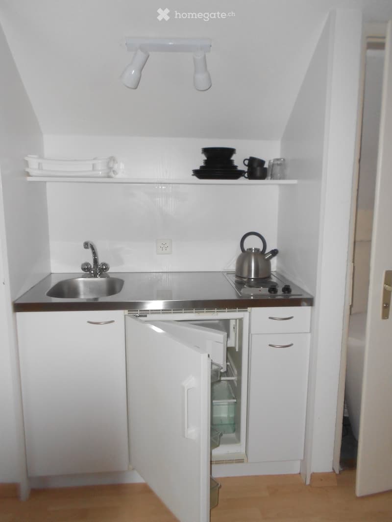Compact kitchen with white cabinets, stainless steel sink, stove, and open refrigerator. Shelves hold dishes and cups.