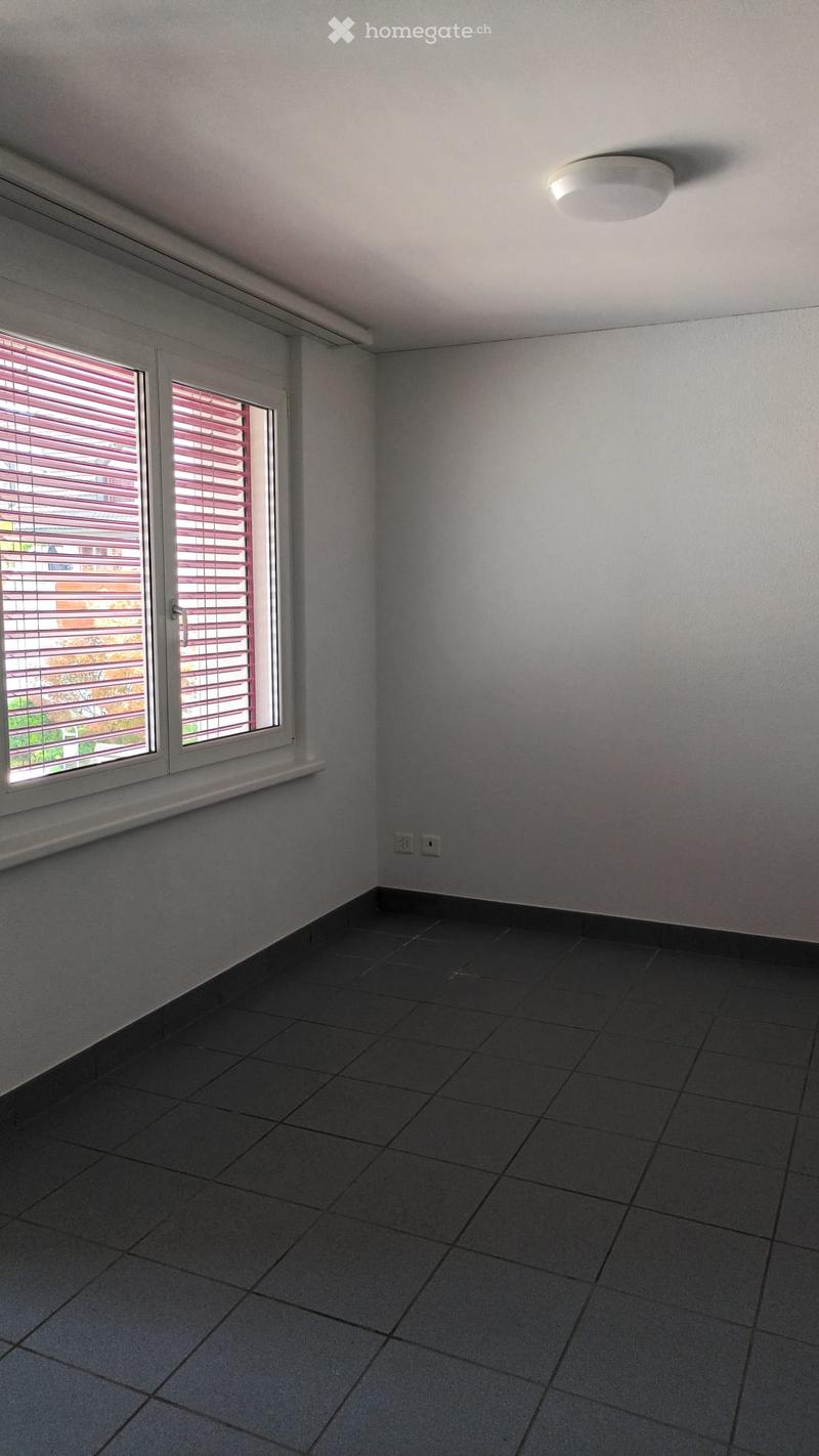 Empty room with grey tiled floor, white walls, and a large window with red blinds. A single ceiling light fixture is visible.