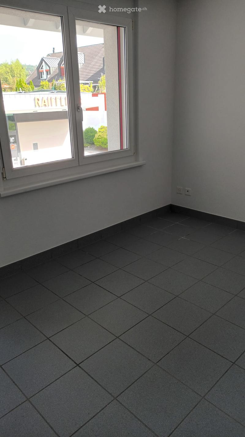 Empty room with grey tiled floor and white walls, featuring a large window with a view of the outside area.