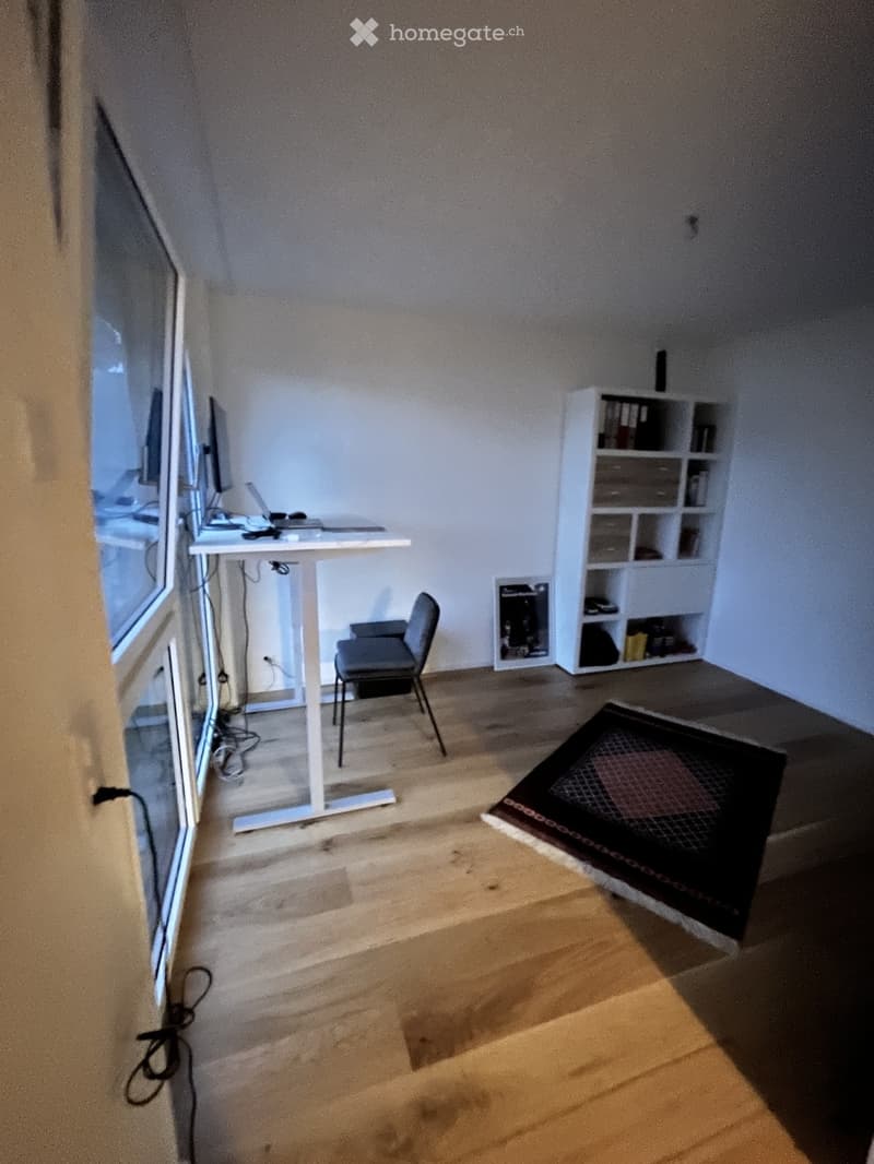 A small, furnished room with a wooden floor, a white desk, a chair, a laptop, a screen, and a white shelf with books and items.