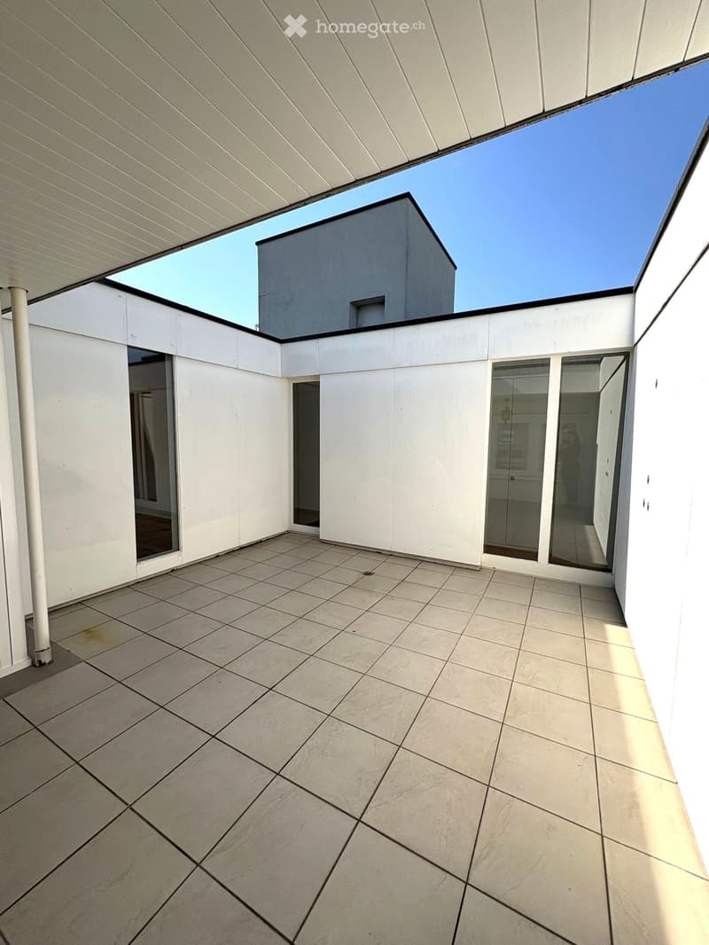 Modern rooftop terrace with tiled flooring, white walls, large glass panels, and a view of a neighboring building.