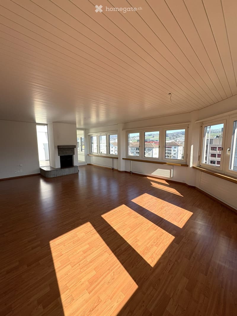 Empty room with wooden flooring, large windows, and a fireplace. Sunlight streams in, casting shadows on the floor.