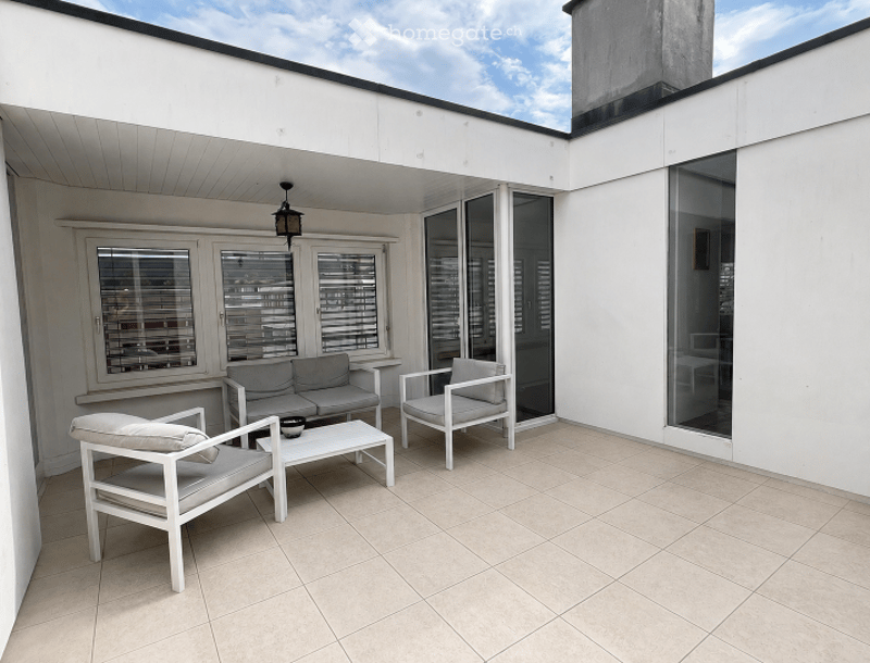 Modern balcony with white furniture, including a sofa, chairs, and a coffee table, large windows, and a hanging lamp.