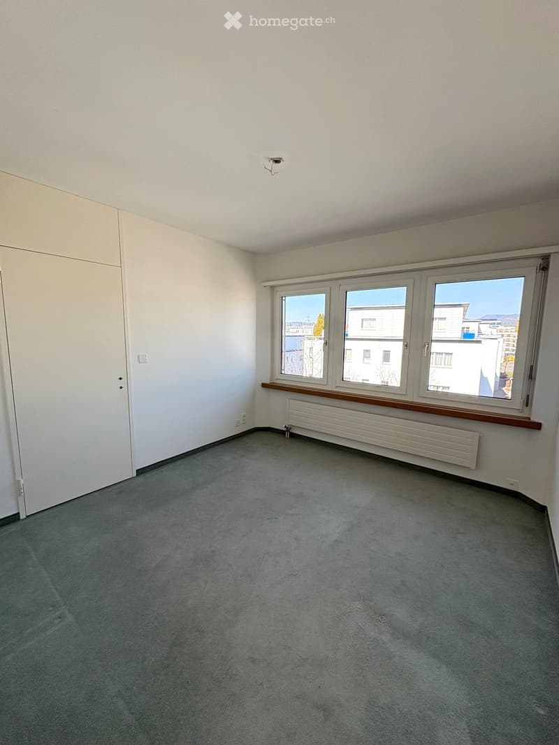 Empty room with grey carpet, white walls, large windows, radiator, and a door.