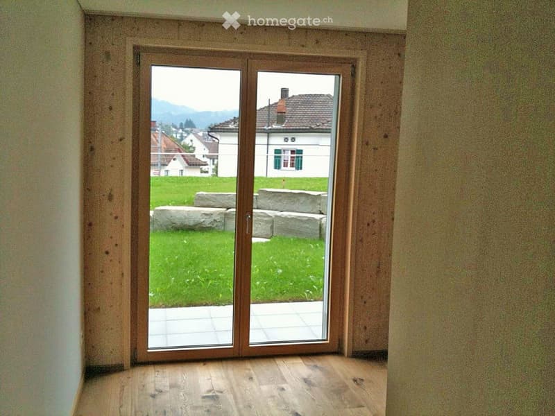 Empty room with wooden flooring and large sliding glass doors opening to a grassy outdoor area with stone benches.