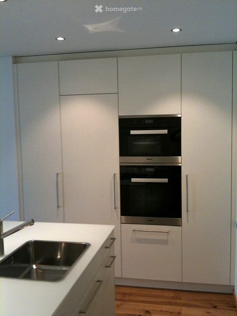 Modern kitchen with white cabinets, stainless steel sink, and double oven. Recessed ceiling lights illuminate the space.