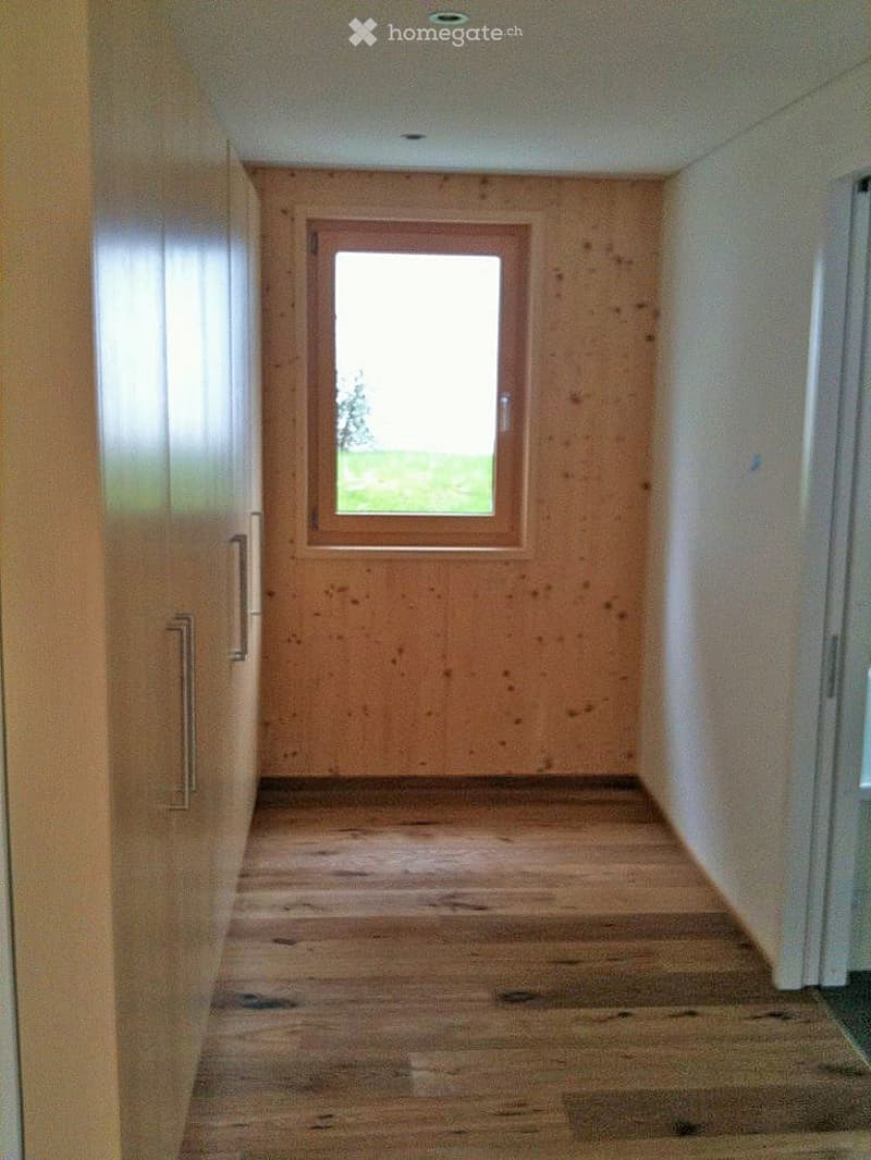 A narrow hallway with wooden flooring, light-colored walls, and a single window at the far end, allowing natural light to enter.