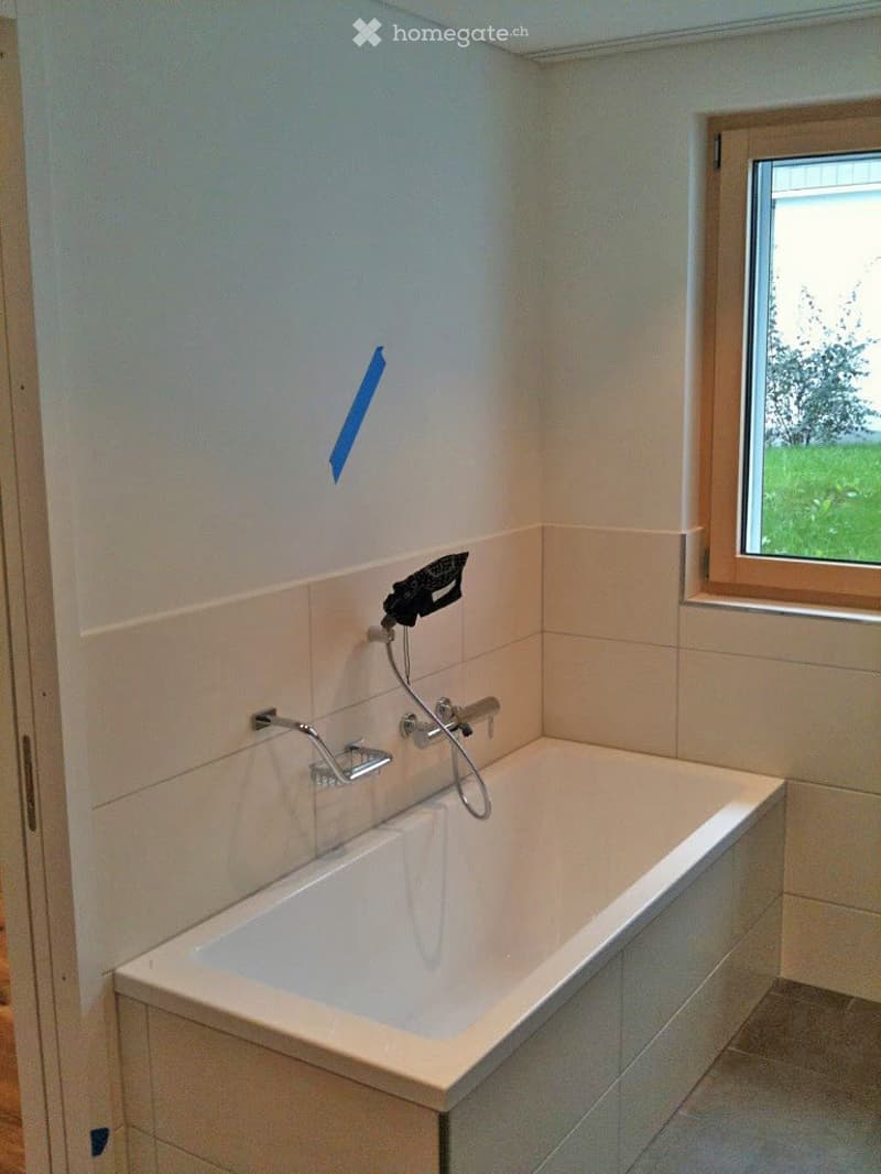 Bathroom with a white bathtub, chrome faucet, and handheld showerhead. Light-colored tiles and a window with a view of greenery.