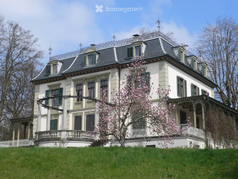 A large, two-story house with white walls, green shutters, and a grey roof. The house features a balcony with ornate railings and columns, surrounded by a well-maintained lawn and blooming pink flowers.