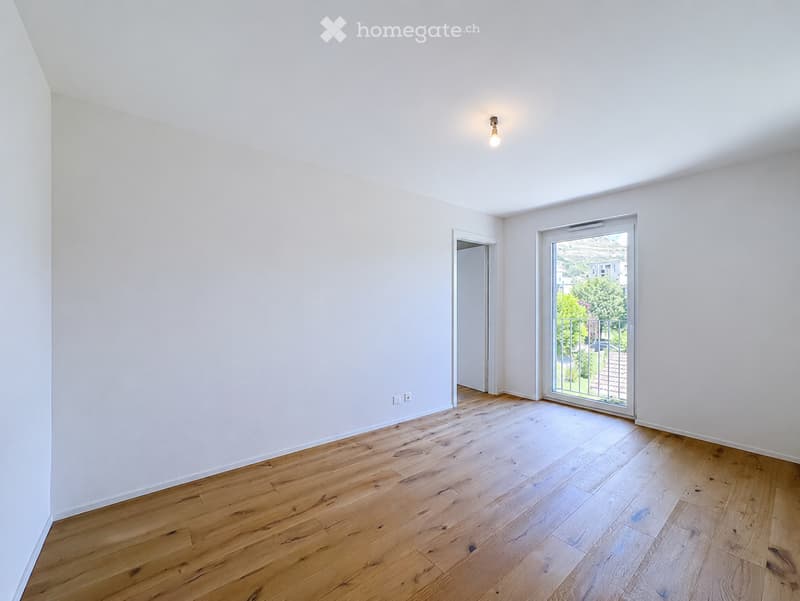 Empty room with light wooden flooring, white walls, and a large sliding glass door leading to a balcony with a view of greenery.