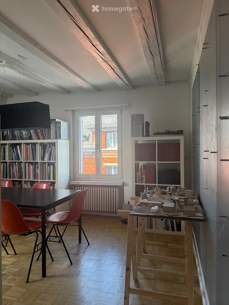 Bright attic room with wooden flooring, large window, and modern furniture including a table, chairs, and bookshelves.