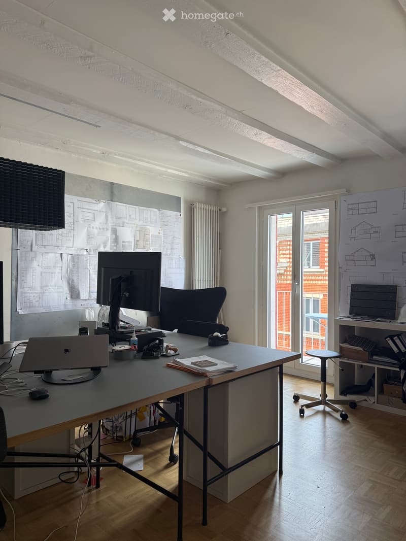 Office space with a large wooden table, laptop, screen, chair, stool, and shelves with papers and drawings.