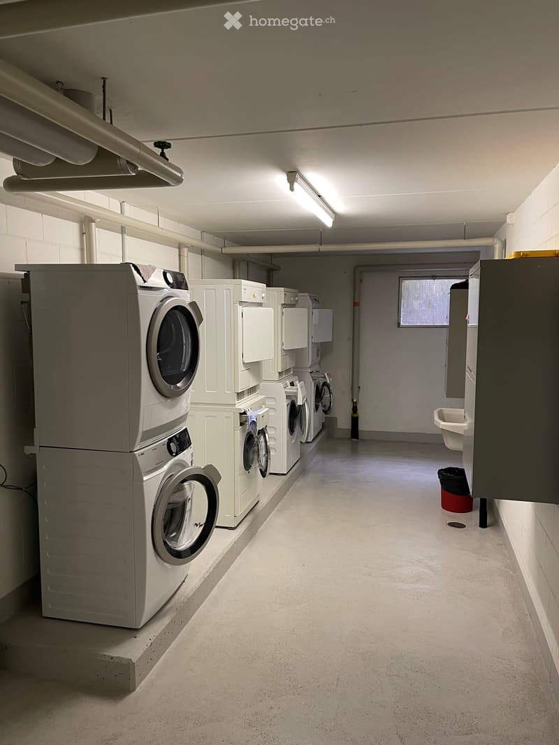A clean, well-lit hallway with multiple stacked washing machines and dryers along one wall, a small sink, and a window at the far end.
