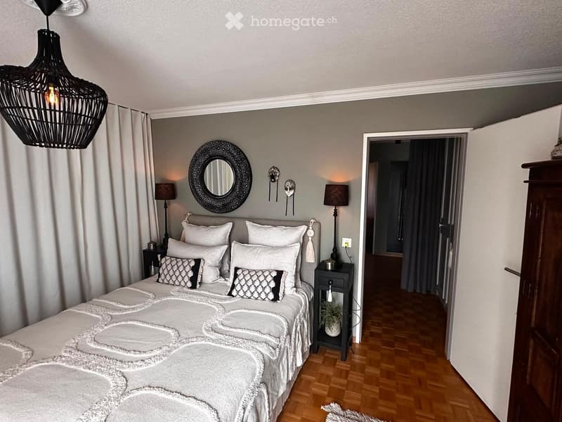Modern bedroom with a large bed, decorative pillows, nightstands, table lamps, a round mirror, and a wicker pendant light.